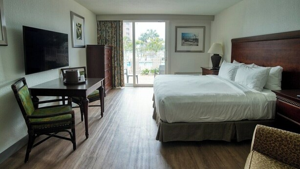 A hotel room with a bed, a desk, a chair, a television, and a window with a view of greenery. - King Standard Room Pool Side (Treasure Island)