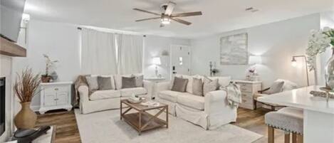 A modern living room with a ceiling fan, white sofas, a coffee table, and a dining area with bar stools.