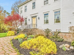 A two-story house with a stone staircase leading to the front door, surrounded by a landscaped garden with various shrubs and a brick pathway. - Modern Country Retreat - 3 Mins to Letchworth - Ideal for Families (Castile)