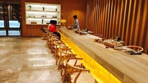Un long comptoir d’accueil avec des chaises en bois, une personne debout derrière, et une étagère avec des objets décoratifs.