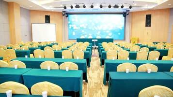 Un salón de conferencias con filas de mesas y sillas, una pantalla grande al frente y un escenario.