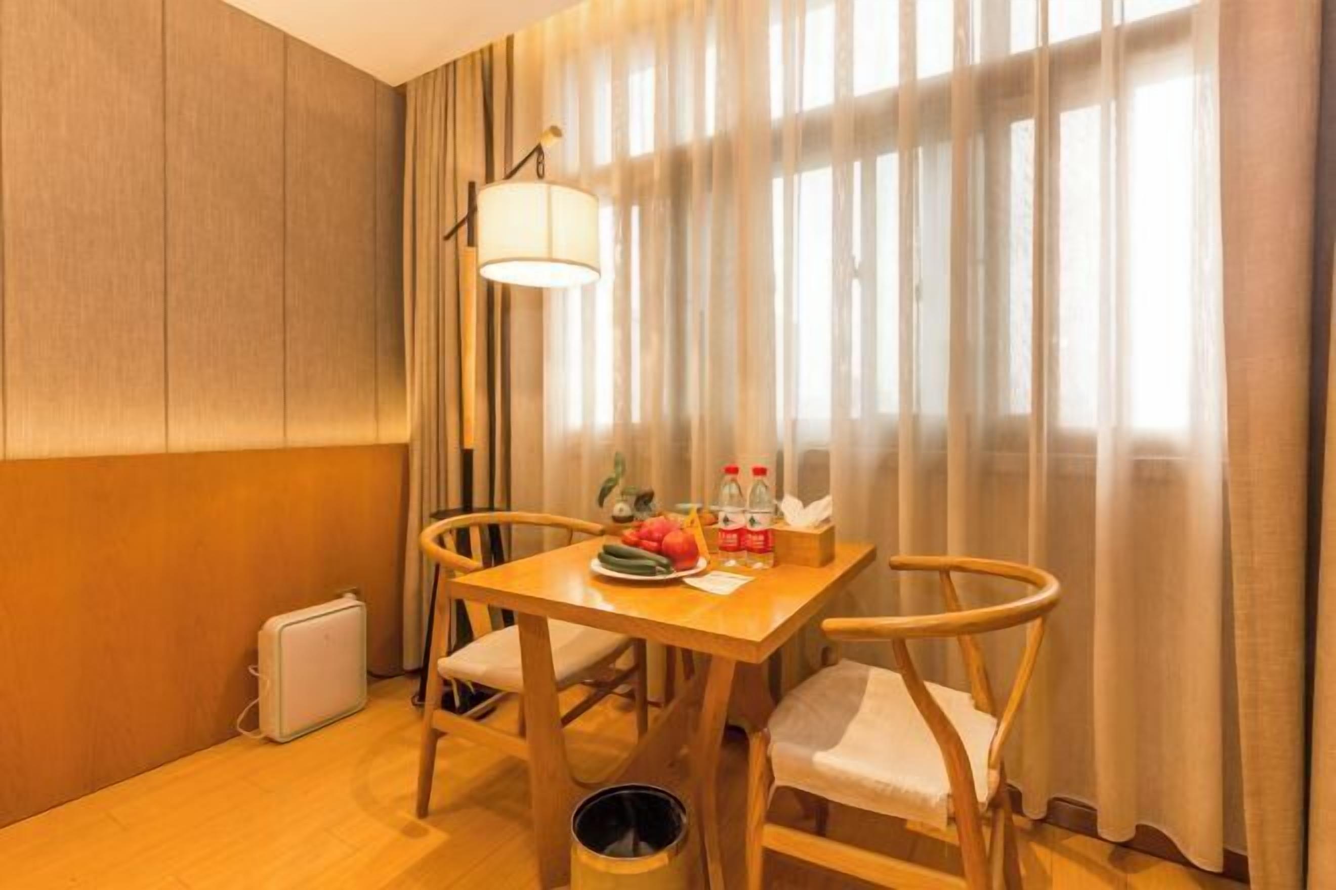 Habitación de hotel con una mesa de madera, dos sillas, una lámpara, un radiador y una ventana con cortinas.