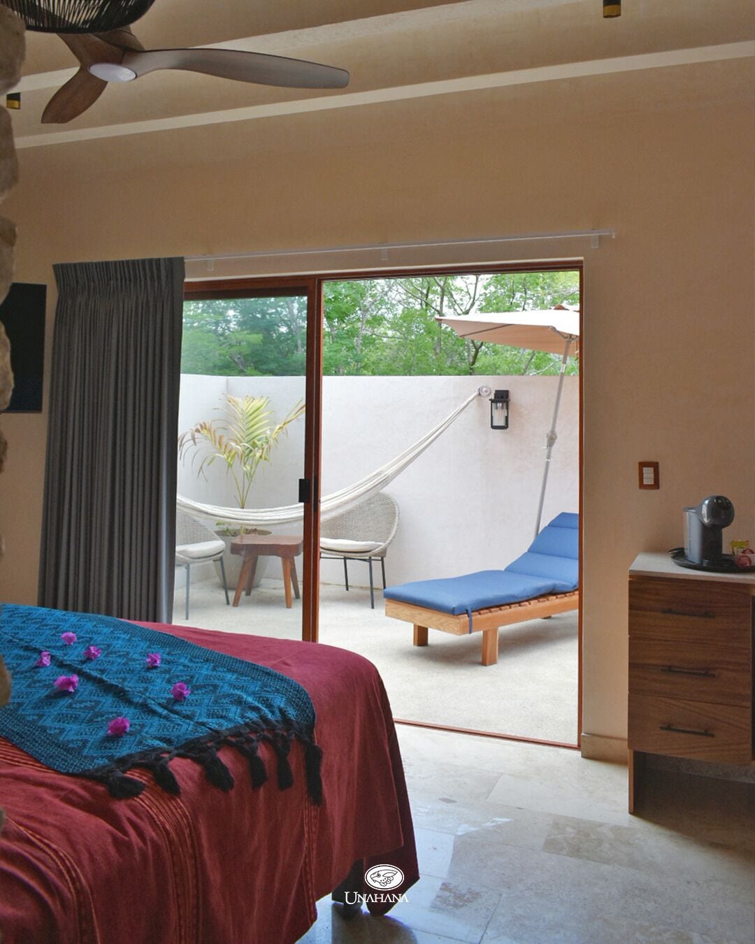 A bedroom with a bed, a desk, a chair, and a hammock outside the sliding glass door.