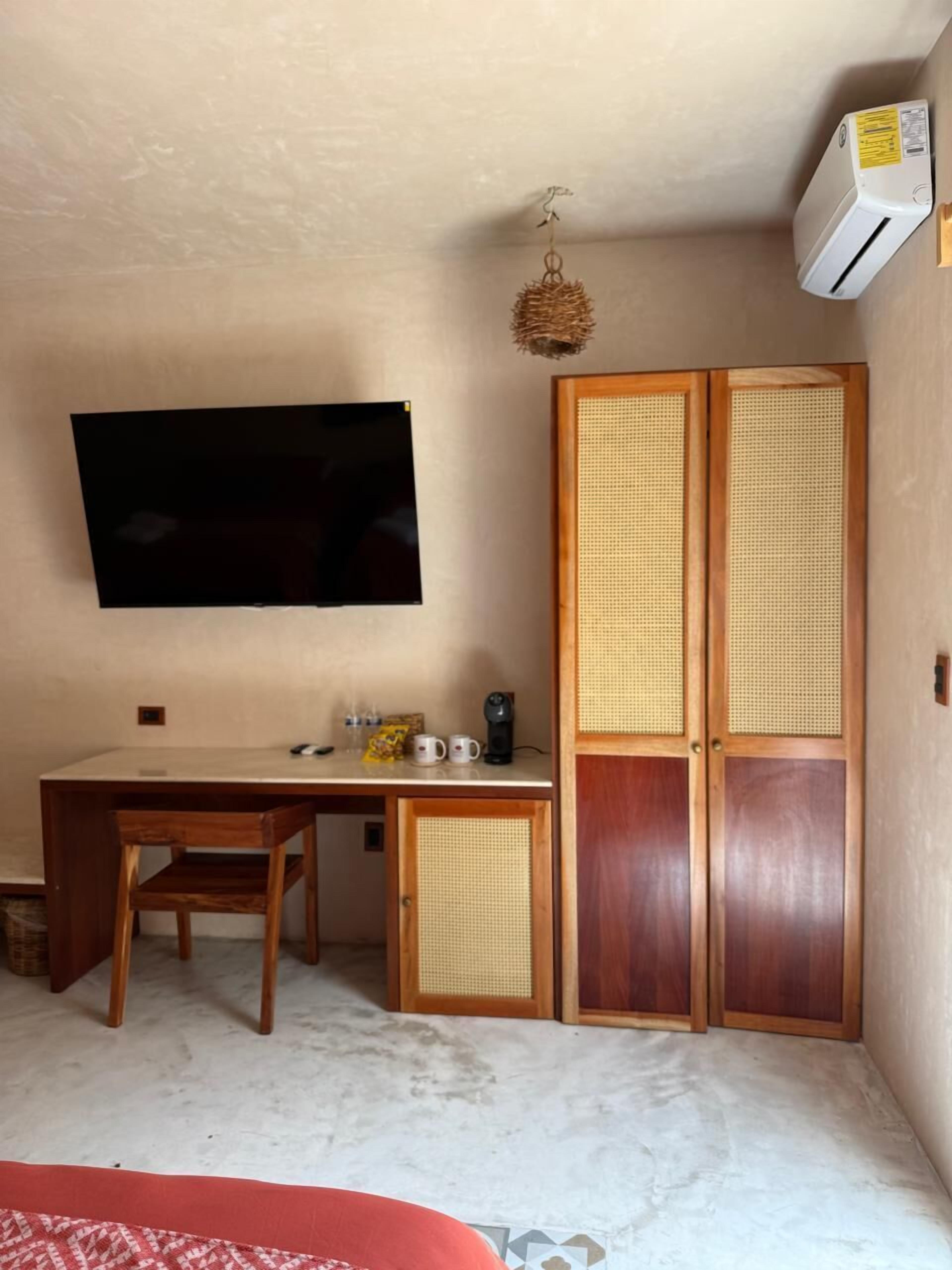 Una habitación con televisión de pantalla plana, un escritorio de madera, una silla, cafetera y armario con puertas de mimbre.