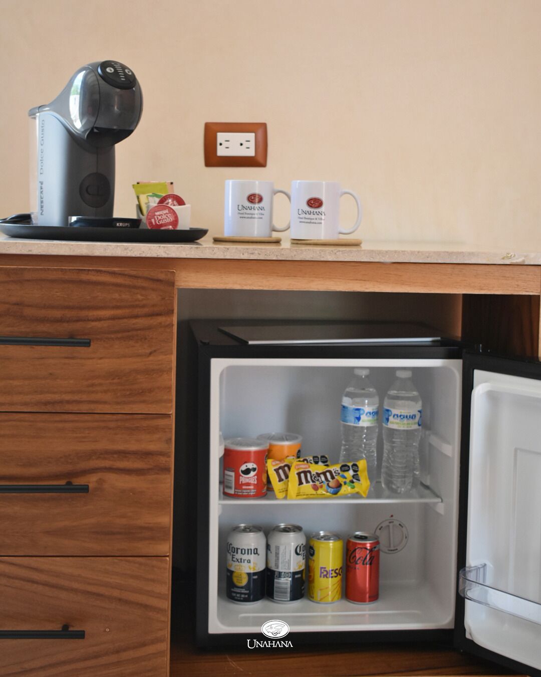 Ein kleiner Kühlschrank mit Getränken und Snacks, eine Kaffeemaschine und eine Steckdose auf der Arbeitsfläche.