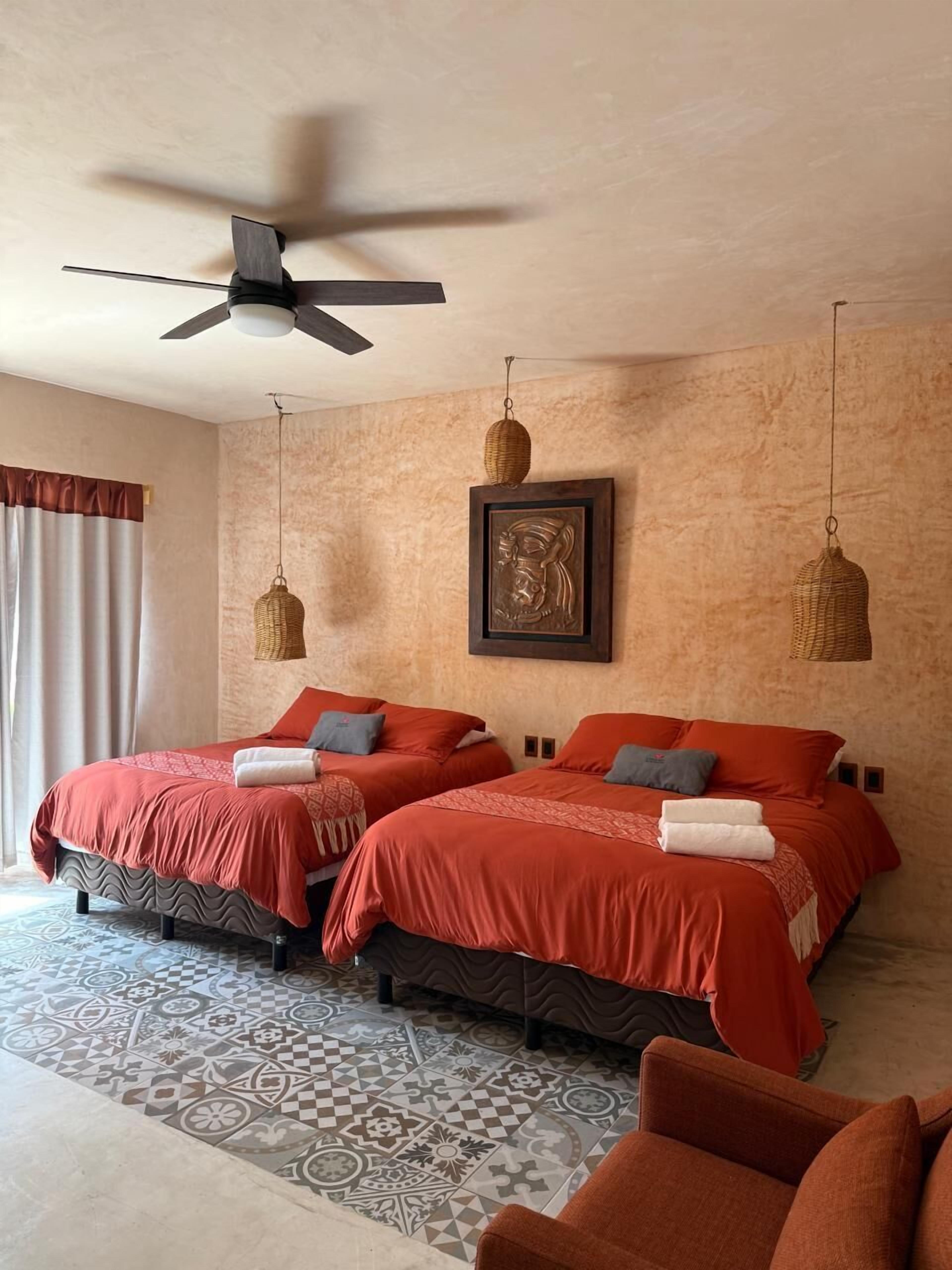 Habitación de hotel con dos camas, un ventilador de techo, un tapete estampado y una obra de arte enmarcada en la pared.