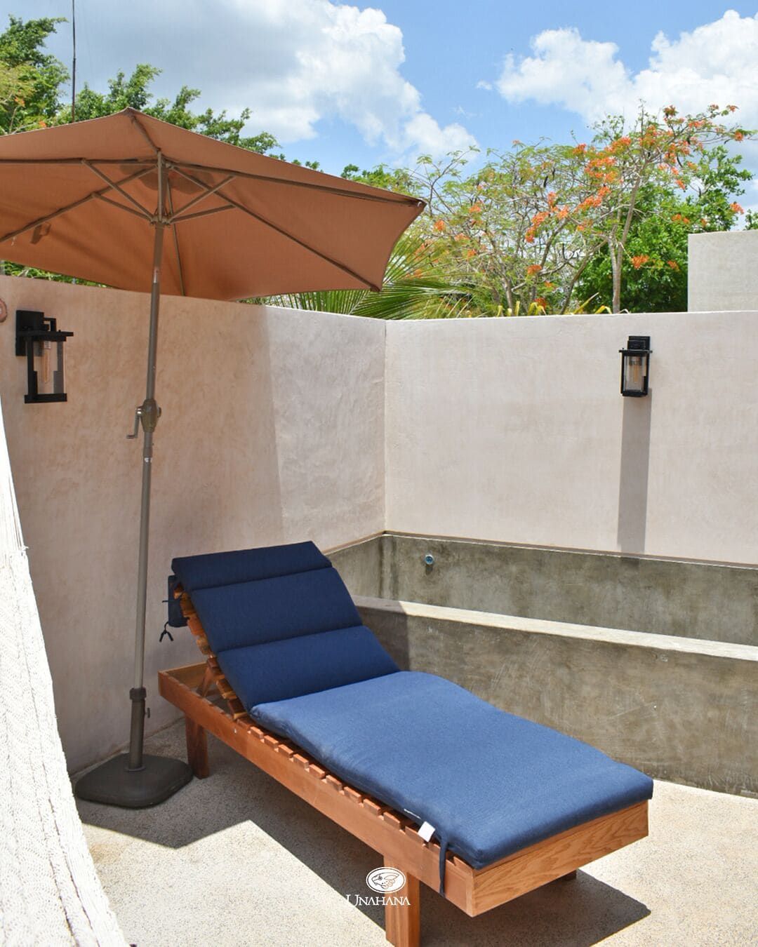 Un fauteuil de jardin en bois avec des coussins bleus, un parasol marron et une lanterne fixée au mur, le tout dans un espace patio extérieur.