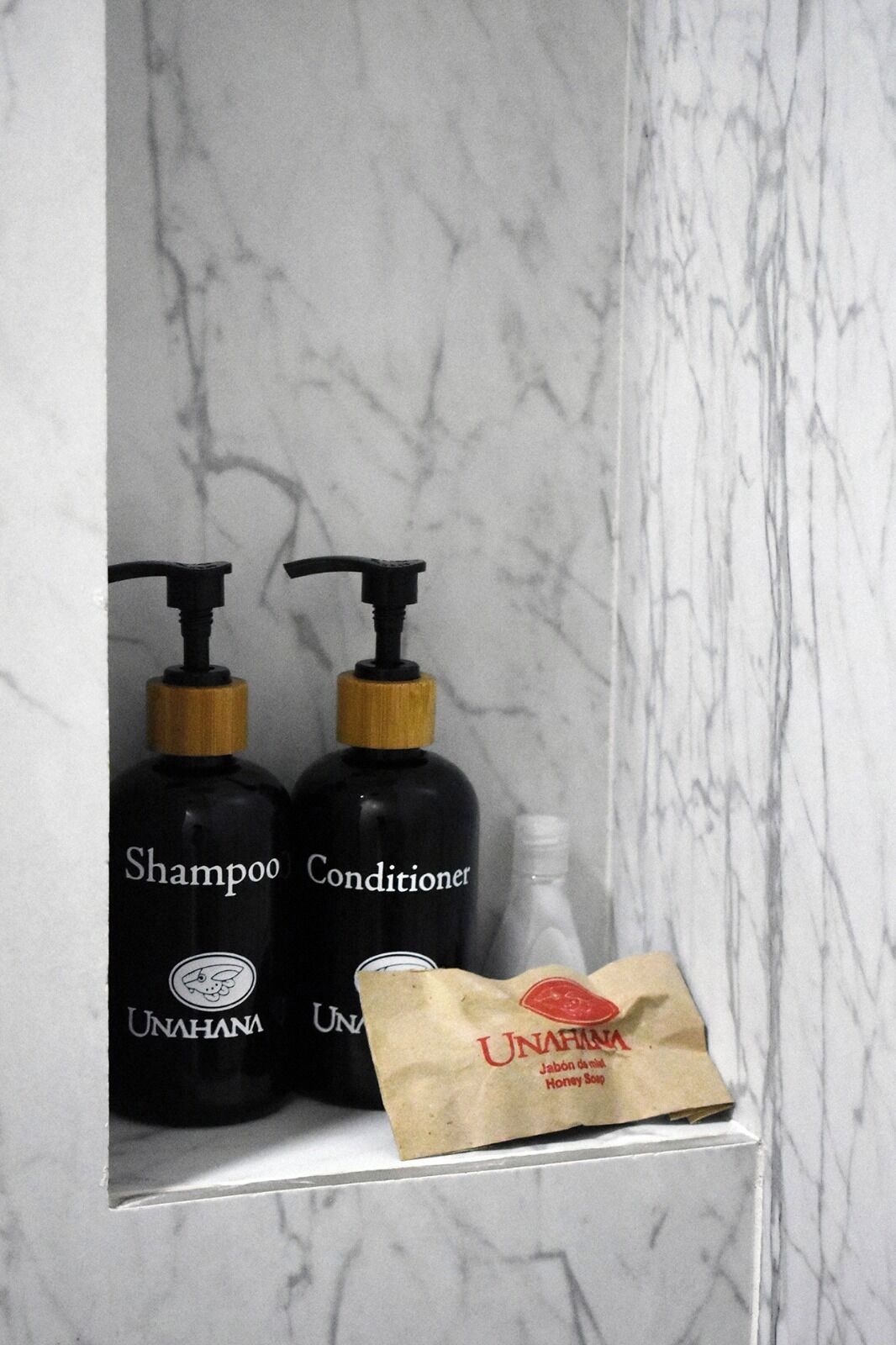 A bathroom shelf with two black bottles labeled 'Shampoo' and 'Conditioner' from 'UNAHANA,' and a brown paper bag with 'UNAHANA' branding.