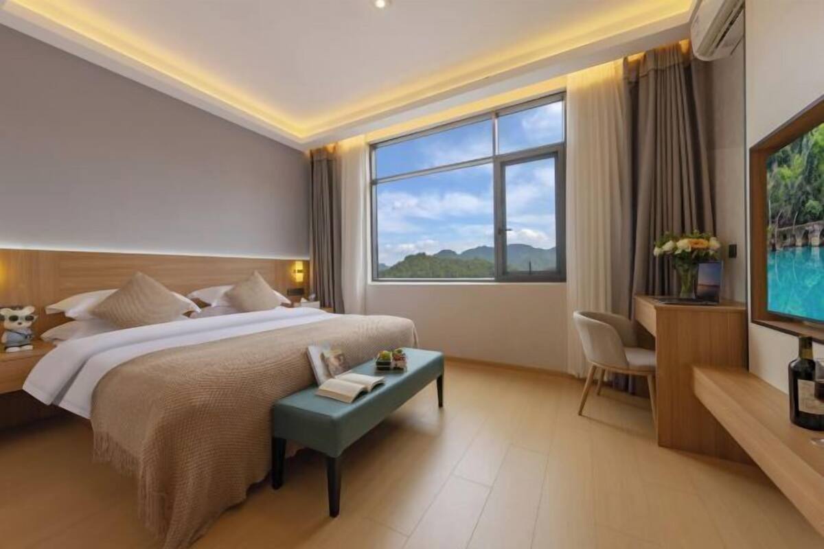A hotel room with a bed, bedside table, chair, TV, and a large window with a view.