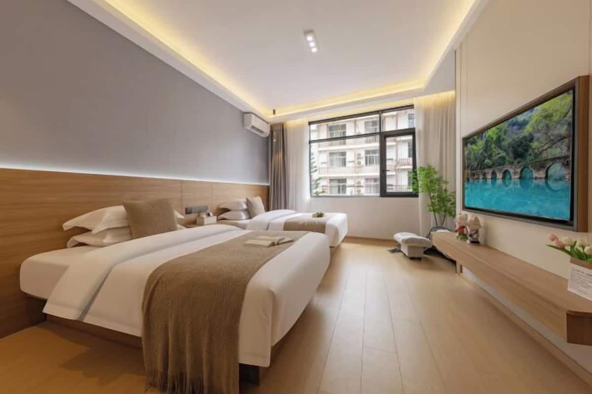 A modern hotel room with two beds, a large window with a city view, a flat-screen TV, and a small seating area.