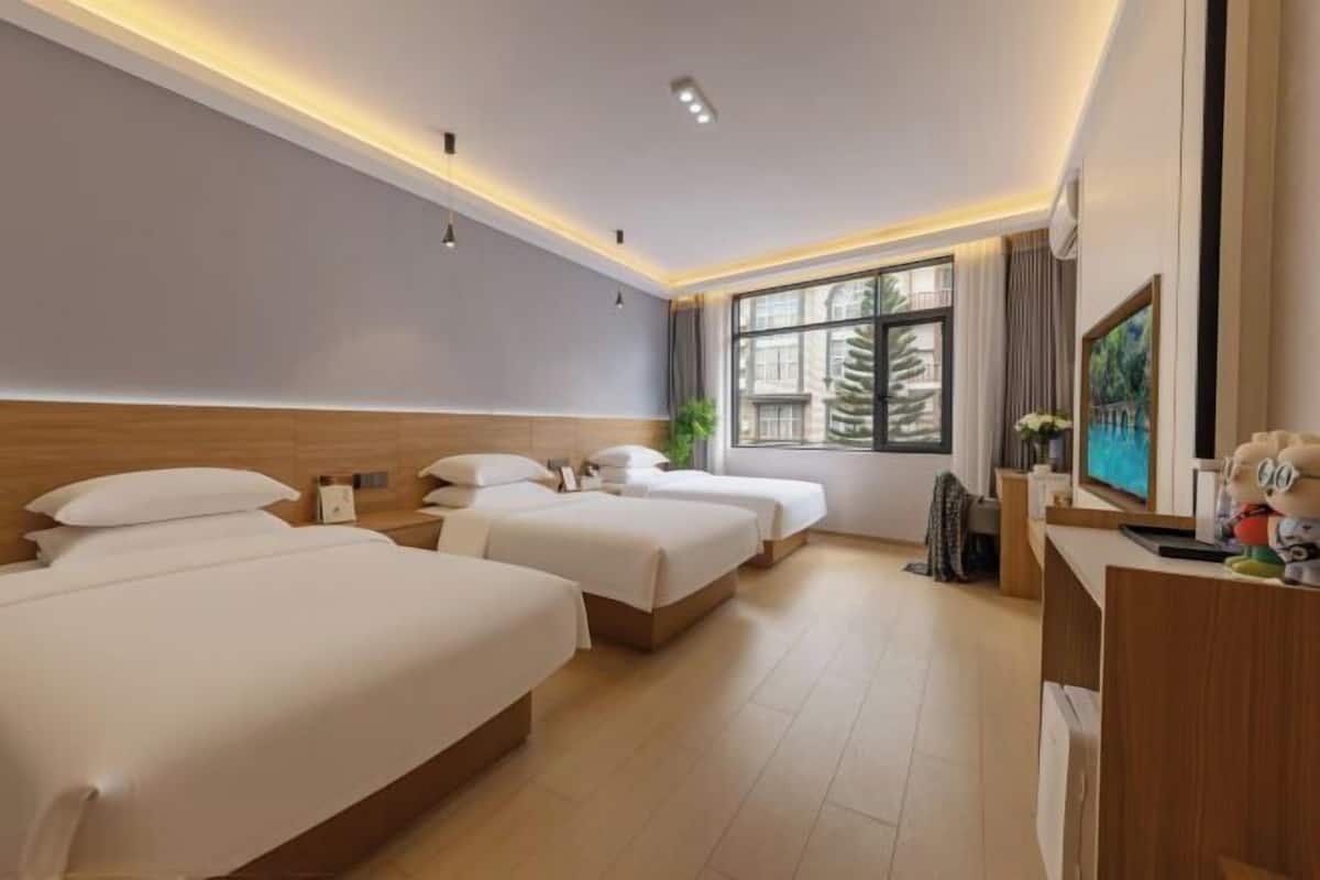 A hotel room with three beds, a desk, a large window, and a plant.