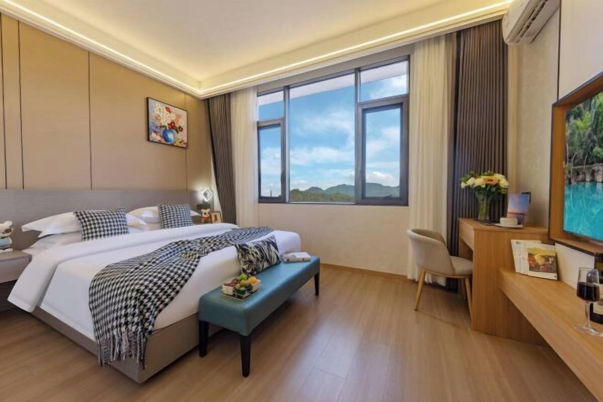 A hotel room with a large bed, a desk, a chair, a window with a view, and a painting on the wall.