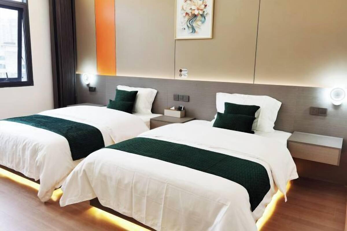 A hotel room with two beds, each with white bedding and green accents. There are bedside tables with lamps, a wall-mounted artwork, and a window with curtains.