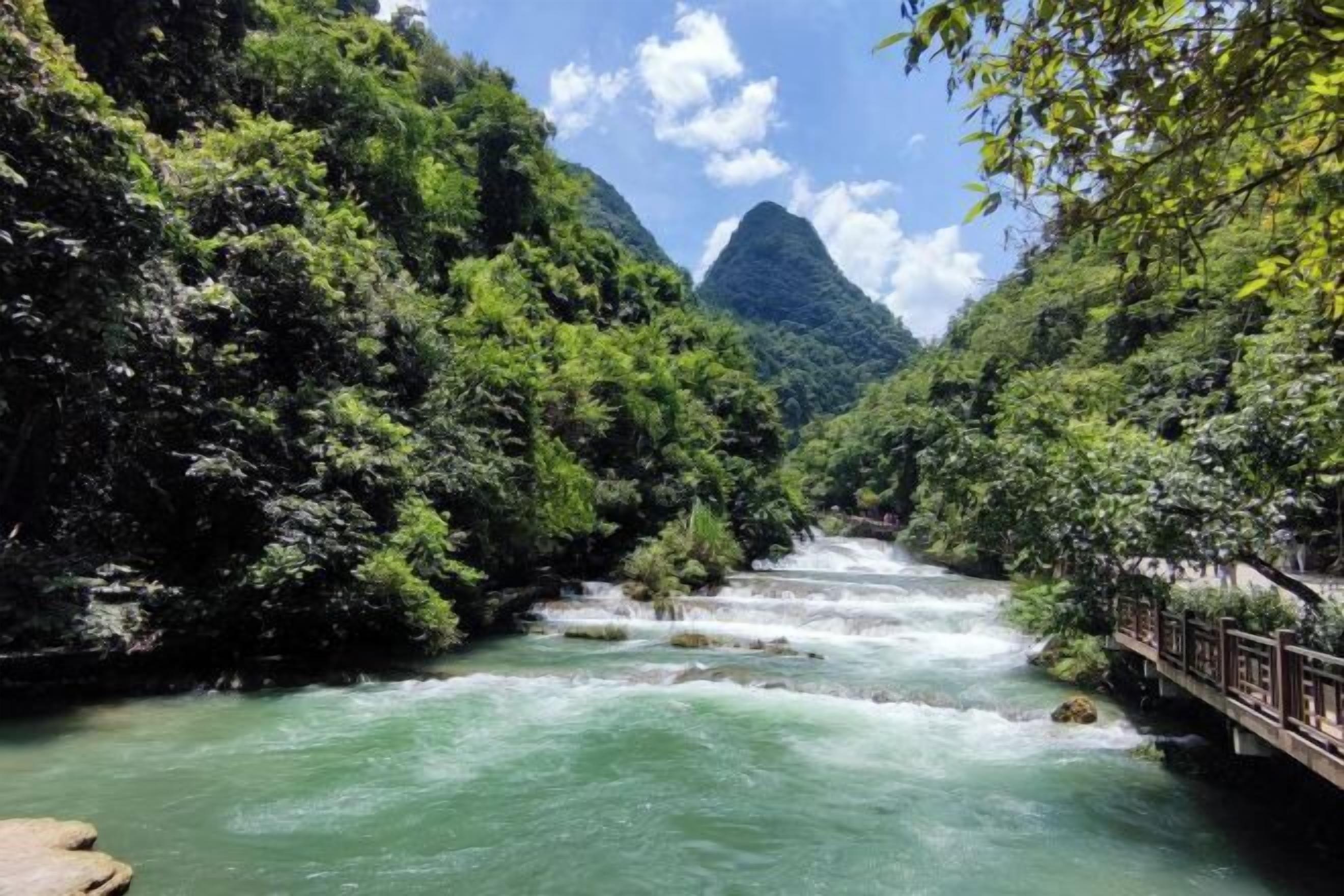 Une rivière serpente à travers une vallée verdoyante et luxuriante, avec un pont et une montagne en arrière-plan.