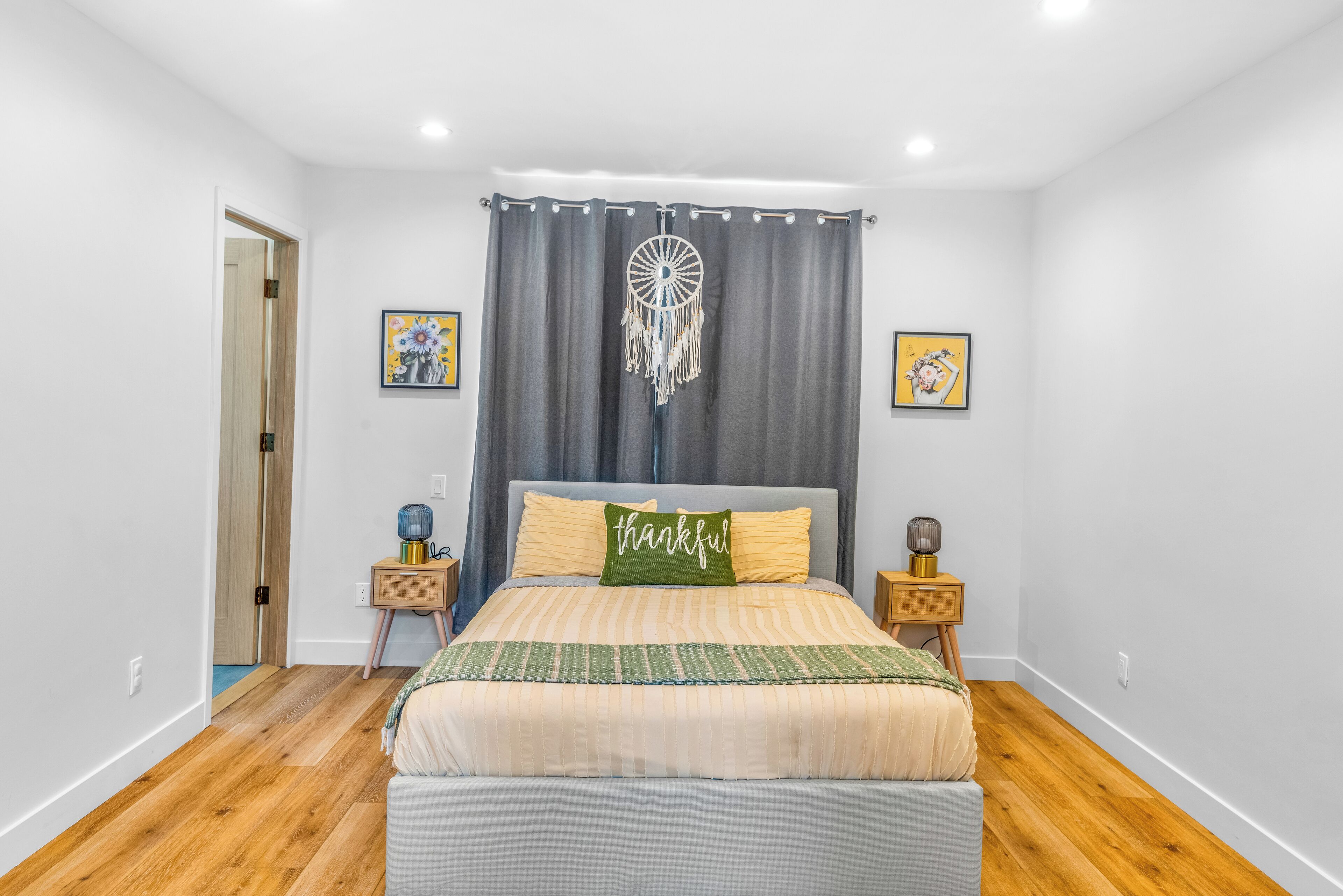 Un dormitorio con una cama, un atrapasueños, dos cuadros enmarcados y una mesita de noche con una lámpara.