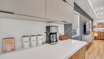 A modern kitchen with a white countertop, coffee maker, sugar and tea containers, and a basket.
