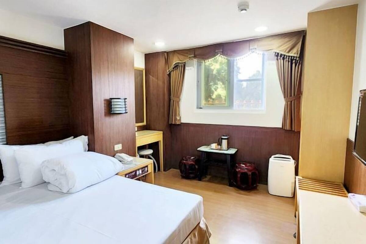 Hotelkamer met een bed, een klein tafeltje met waterkoker, een stoel, een bureau, een spiegel, een raam met gordijnen en een wandlamp.
