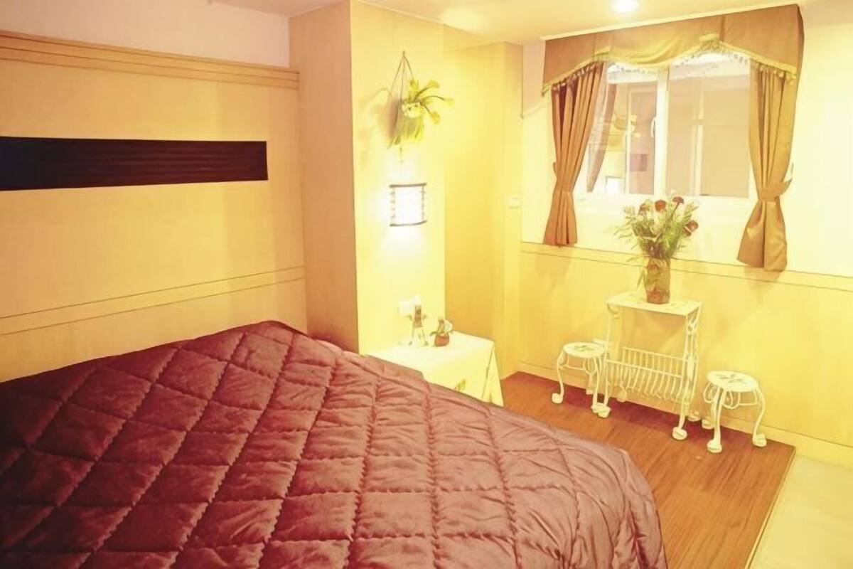 Een slaapkamer met een groot bed, nachtkastje, lamp, raam met gordijnen en een klein krukje.