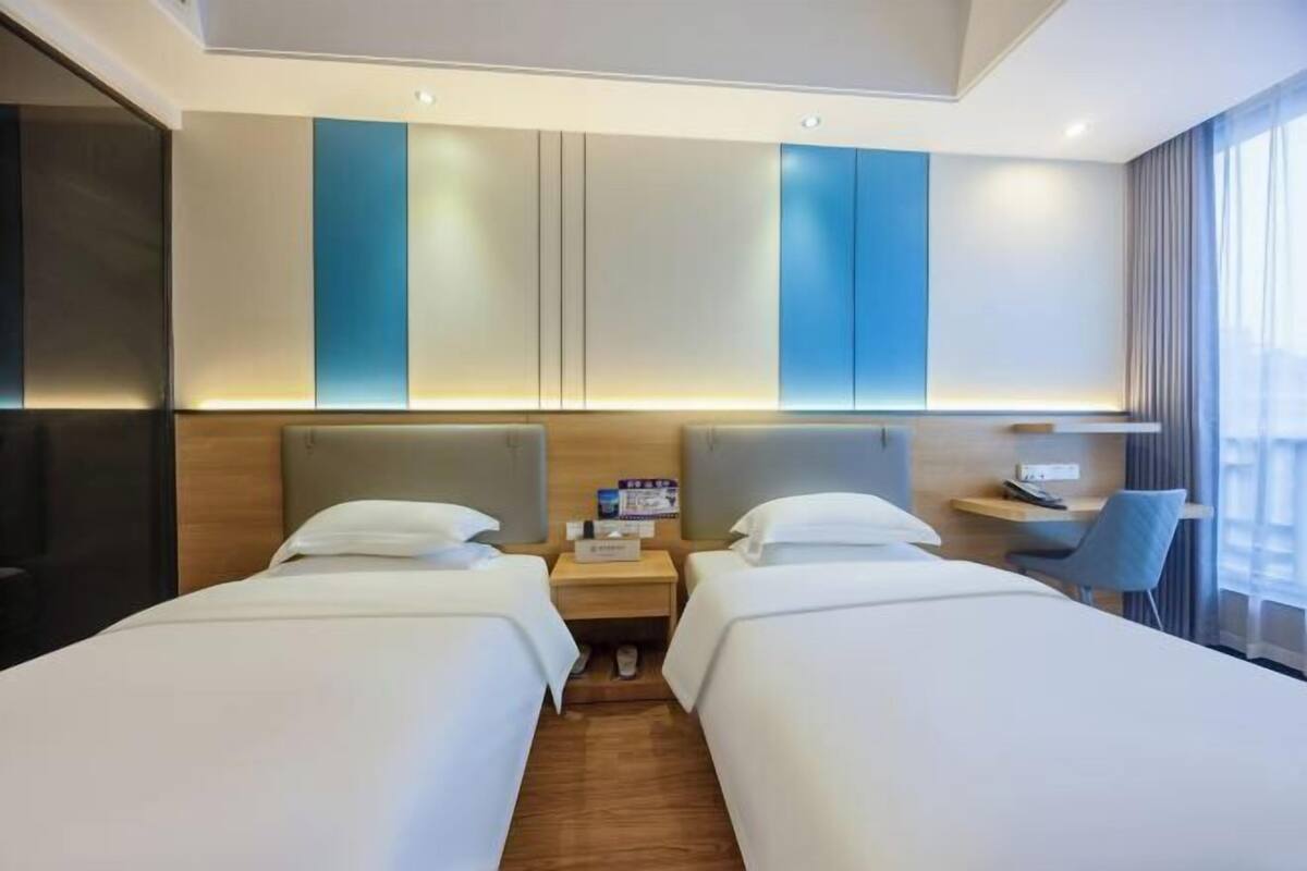 Ein Hotelzimmer mit zwei Einzelbetten, einem kleinen Schreibtisch, einem Stuhl und einem Nachttisch mit einer Lampe.