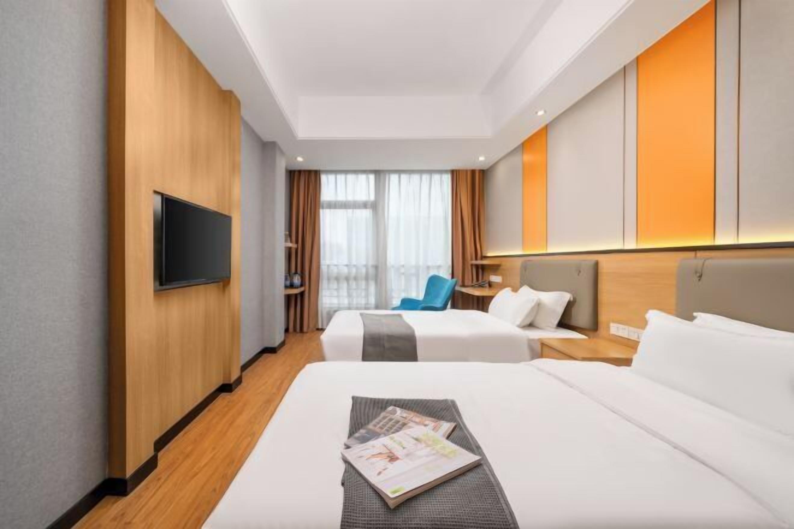 A hotel room with two beds, a flat-screen TV mounted on the wall, a wooden accent wall, and a large window with curtains.