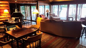Un salon chaleureux avec une table en bois, des chaises, un canapé et une table basse. Il y a deux lampes sur la table et une grande fenêtre donnant sur les arbres.