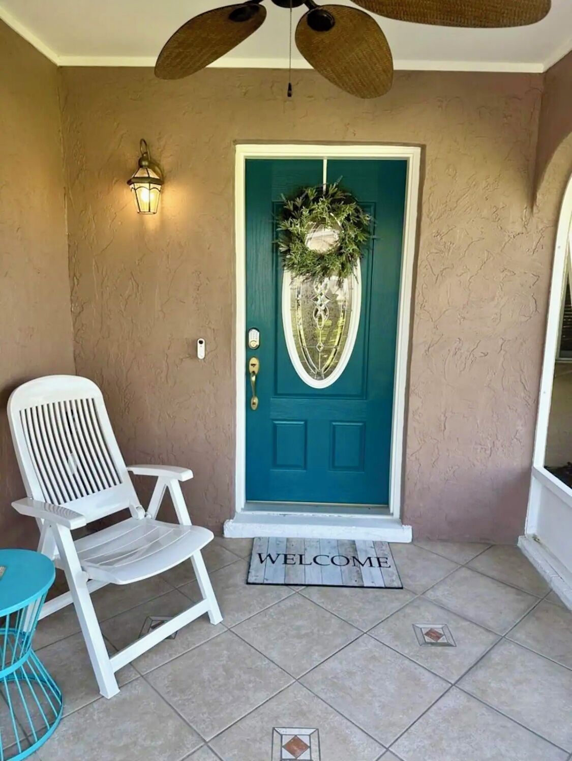 Una puerta frontal color turquesa con un adorno de corona, una silla blanca y un felpudo con la palabra 'WELCOME'.