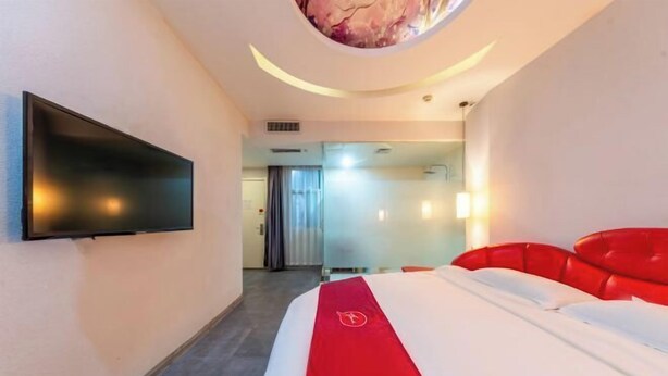 A hotel room with a bed, a flat-screen TV mounted on the wall, and a red headboard.