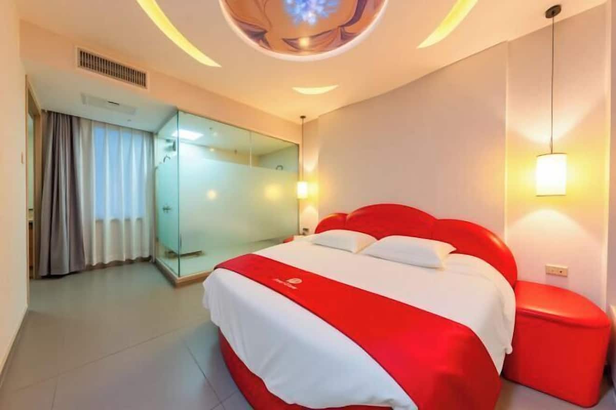 Une chambre d’hôtel avec un lit, une cloison en verre, un pouf rouge et un luminaire au plafond.