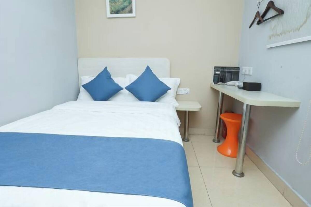 A bedroom with a bed, pillows, a bedside table, a nightstand, a small orange stool, and a wall-mounted telephone.
