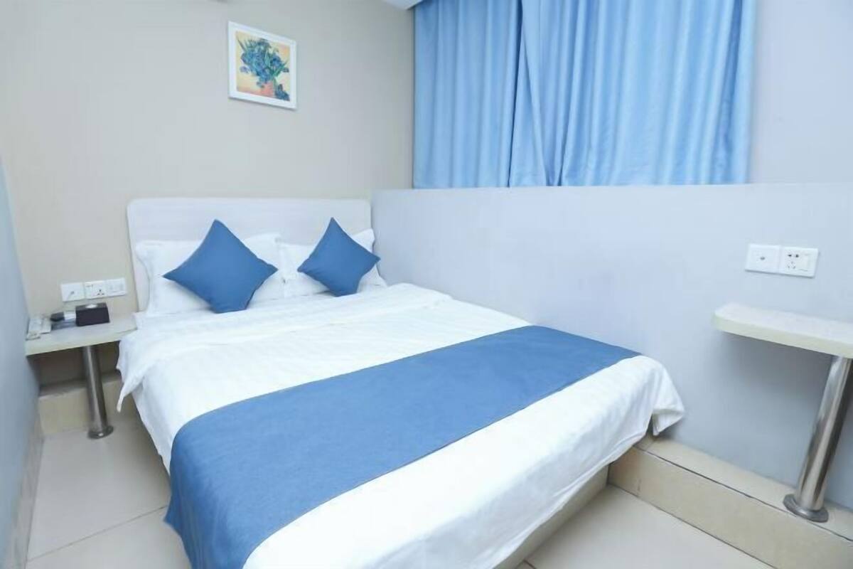A hotel room with a bed, a nightstand, a bedside table, a wall-mounted power outlet, and a window with curtains.