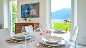 A dining table set with plates, glasses, and cutlery, with a view of a lake and mountains through large windows. - Luxury Central Suite Garden in Lugano (Lugano)