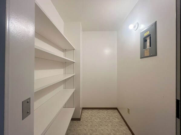 A narrow hallway with white walls and a patterned floor. Shelves are mounted on the left wall, and a light fixture is visible on the right. - WorkStay Lodging - Spy Hill, SK - Near Esterhazy, Rocanville & Potash Mine Sites (Spy Hill)
