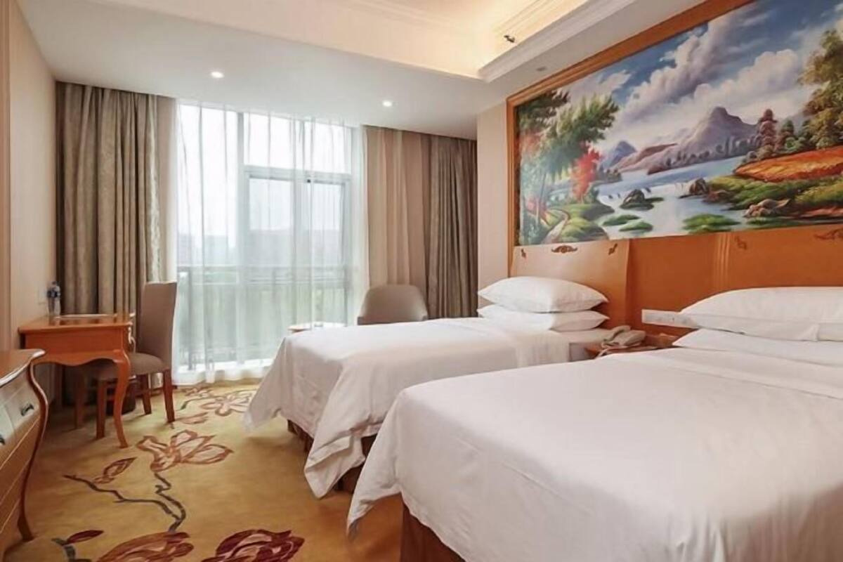 A hotel room with two beds, a desk, and a chair. There is a painting on the wall and a window with curtains.