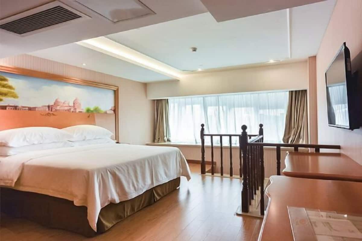 A hotel room with a bed, a desk, a painting of a cityscape, and a window with curtains.
