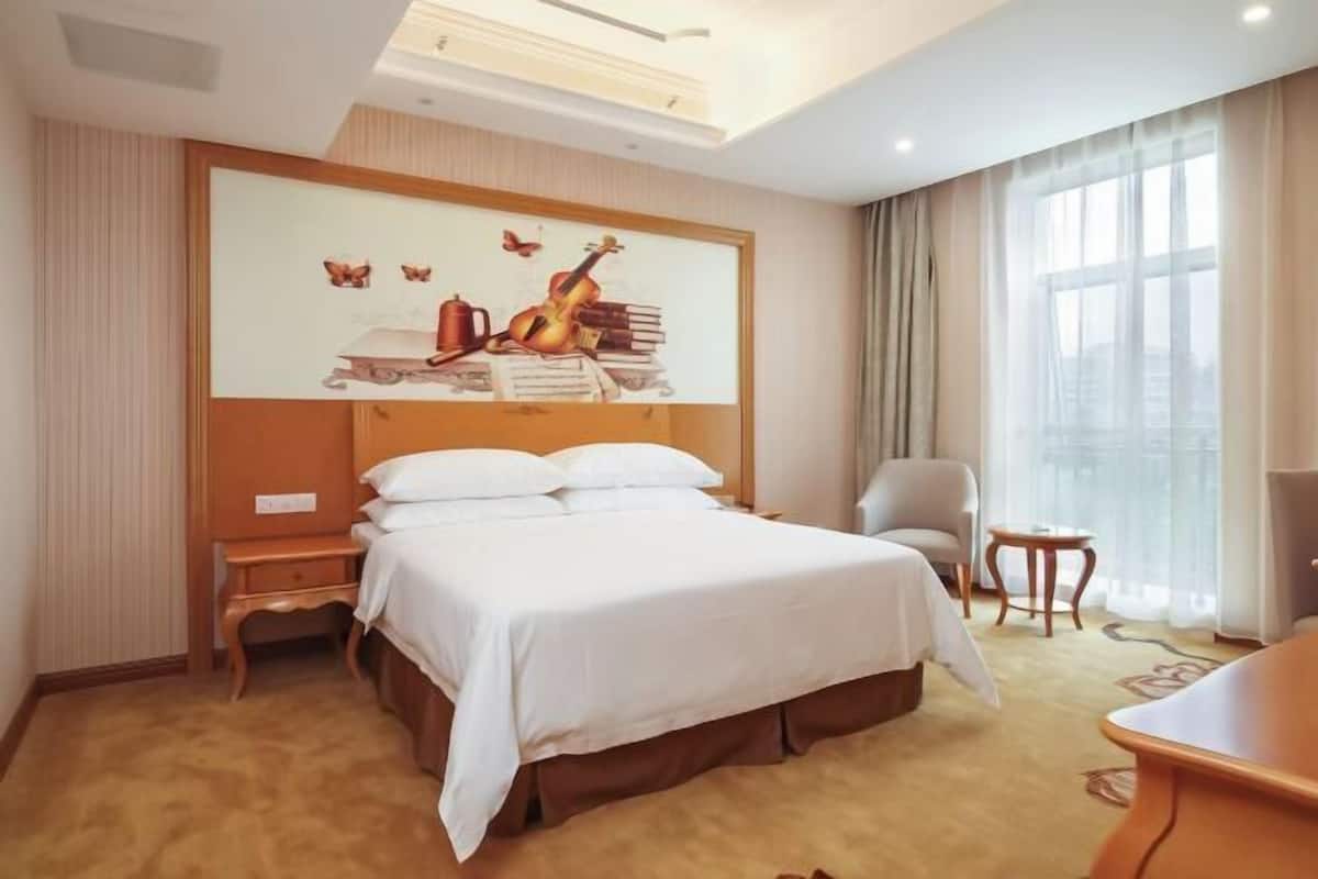 A hotel room with a large bed, a desk, a chair, and a large window.