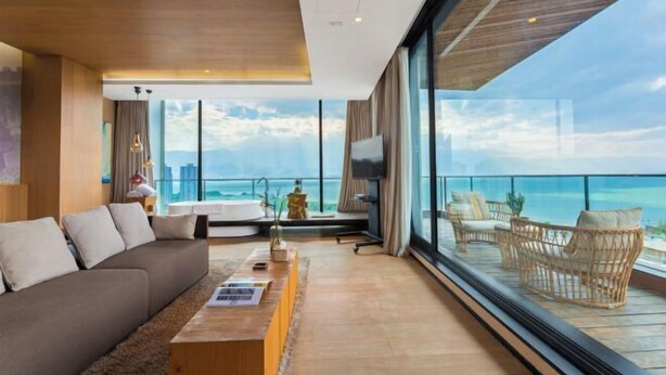 A modern living room with a large sofa, a coffee table, and a TV. There is a balcony with wicker furniture and a view of the ocean. - Dali Hidden in the Clouds Lake View Hotel (Dali City)