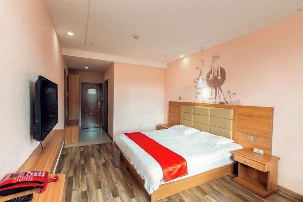 A hotel room with a bed, bedside tables, a TV, and a hallway leading to another room.