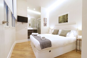 Une chambre moderne avec un grand lit, un téléviseur à écran plat fixé au mur, une salle de bain avec un miroir et un tableau encadré au mur.