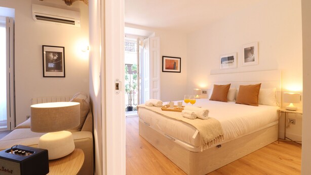 A well-lit bedroom with a bed, bedside table, lamp, and a small table with a speaker. - Reina Sofia Suite II (Madrid)