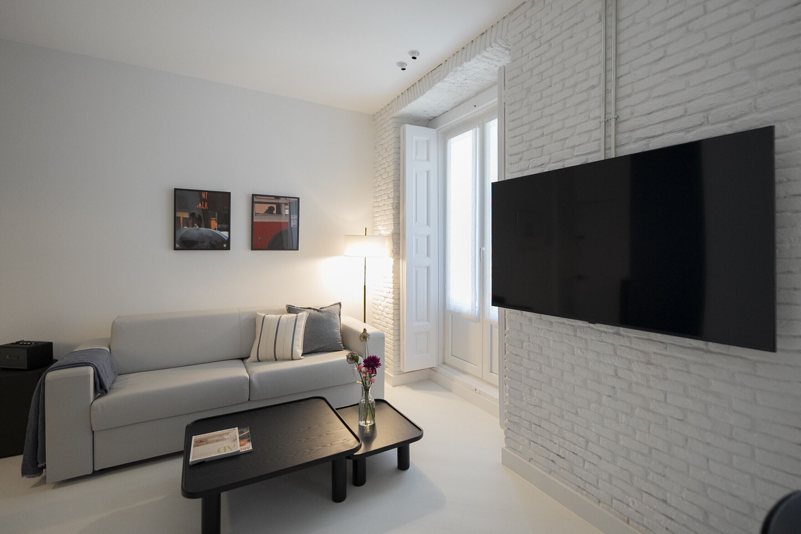 A modern living room with a white brick wall, a flat-screen TV, a grey sofa, a black coffee table, and two framed pictures on the wall.