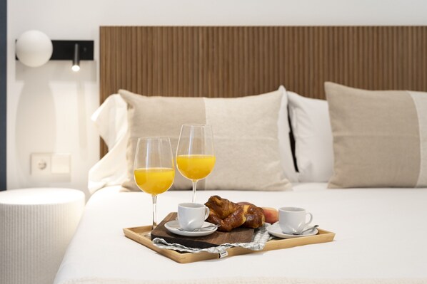 A neatly made bed with a tray of breakfast including croissants, orange juice, and coffee, in a modern hotel room with a wooden headboard and white bedding.
