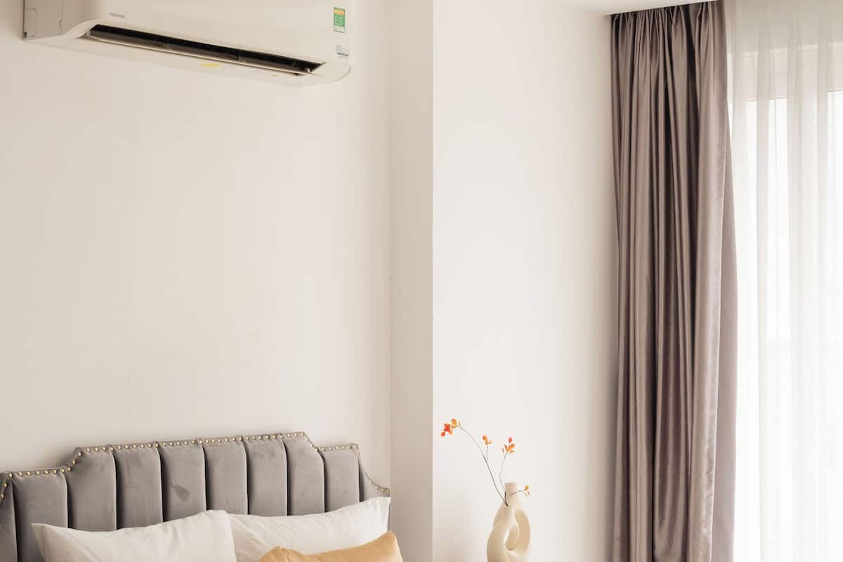 A modern hotel room with a neatly made bed, a bedside table with a vase and books, and a large window with curtains.