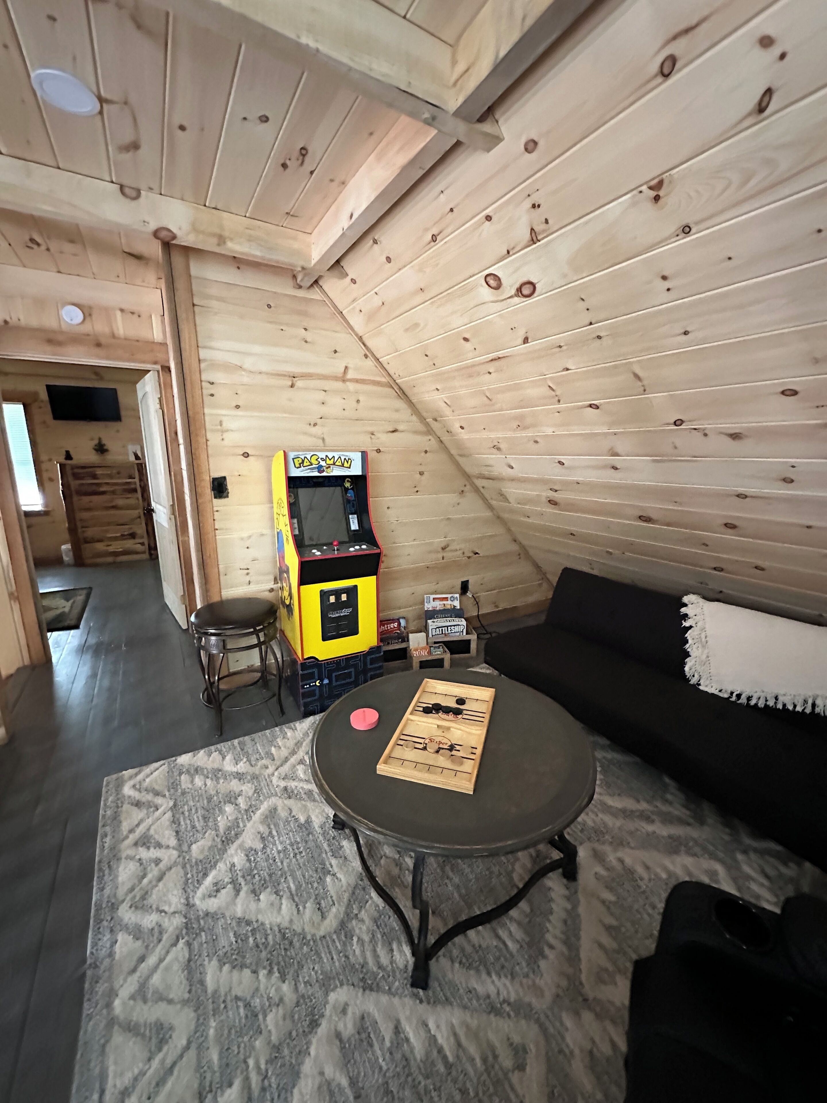 A cozy attic room with a wooden sloped ceiling, a small round table with a board game, a black sofa, and a Pac-Man arcade machine.
