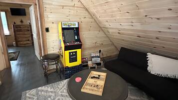 A cozy attic room with a wooden sloped ceiling, a small round table with a board game, a black sofa, and a Pac-Man arcade machine.