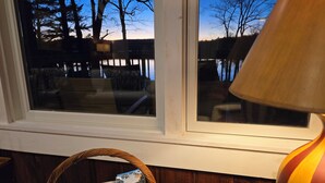 Ein Fenster mit Blick auf Bäume und einen See, ein Korbwäsche mit einem Buch und eine Lampe auf einem Tisch.