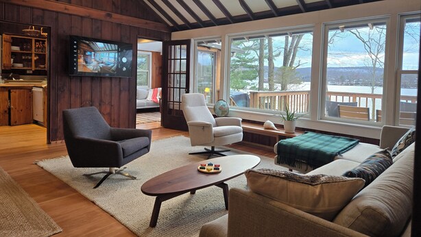 Ein gemütliches Wohnzimmer mit Holzwänden, einem großen Fenster mit Blick auf eine Terrasse und Bäume, einem an der Wand montierten Fernseher und einer komfortablen Sitzecke mit einer Couch und Sesseln.