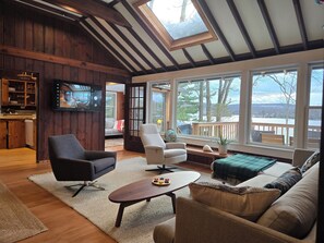 Una sala de estar acogedora con paredes de madera, un ventanal con vistas a una terraza y árboles, un televisor montado en la pared y un rincón de estar cómodo con un sofá y sillones.