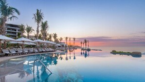 Ein Infinity-Pool mit Liegestühlen und Sonnenschirmen, Palmen und Blick auf den Sonnenuntergang über dem Wasser.