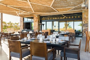 Restaurante com área externa, mesas postas com pratos, copos e talheres, e vista para palmeiras e paisagem desértica.