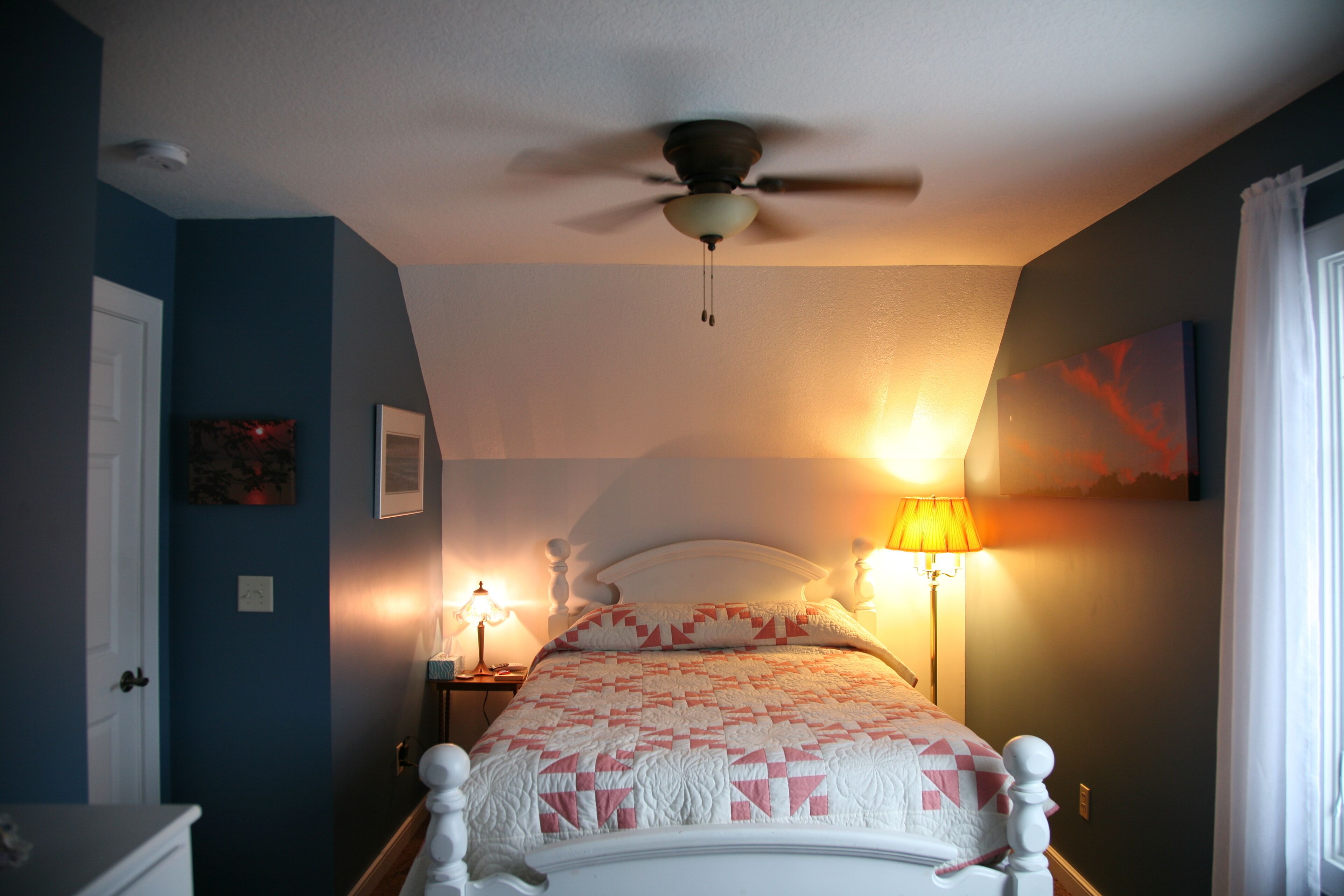 Un dormitorio con cama, ventilador de techo, mesita de noche con lámpara y un cuadro en la pared.